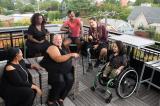 Overhead shot of six disabled people of color at a rooftop deck party. An Indigenous Two-Spirit person with a prosthetic leg smiles directly at the camera and gives a thumbs up while everyone else is engaged in conversation. 