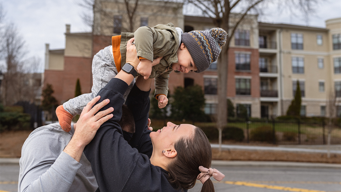 photo of a woman laughing while playfully lifting her one year old toddler son up in the air as her husband affectionately watches