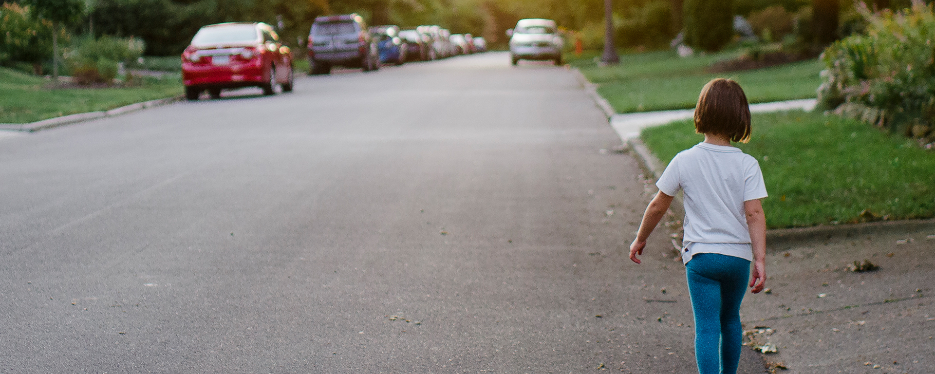 A child walks down the street, back to the camera, lit by the early summer sun.