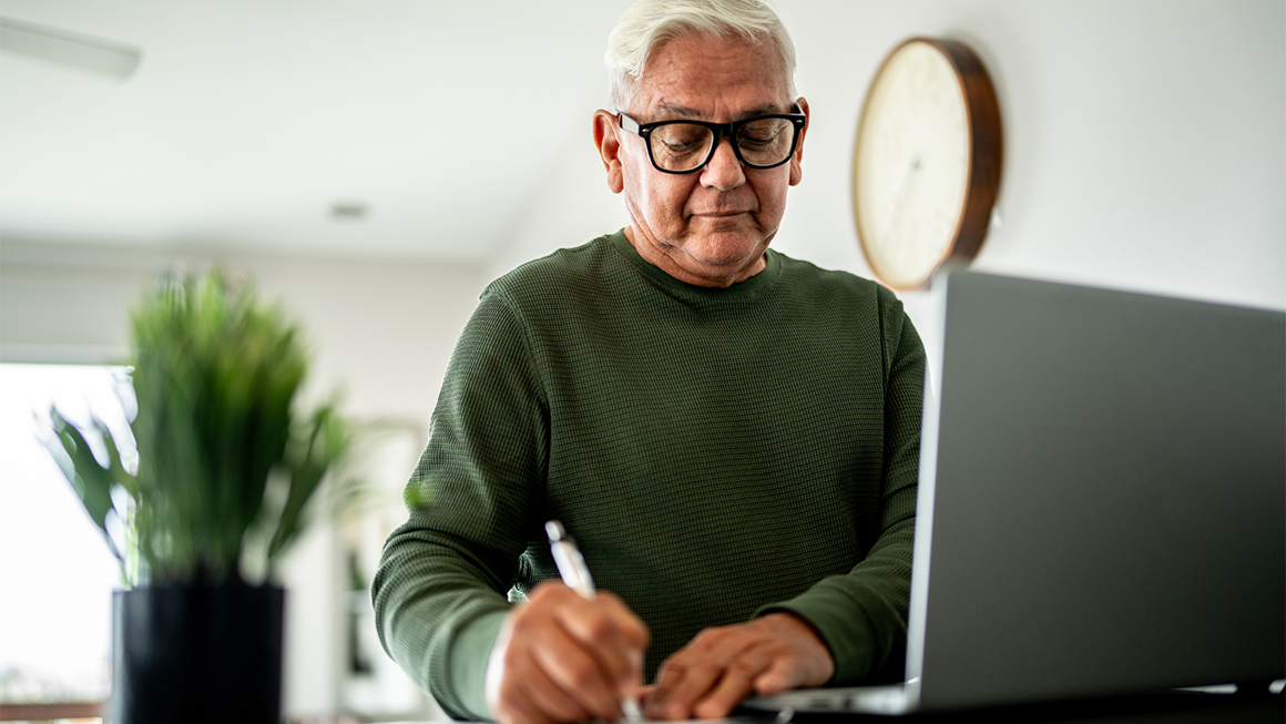 Image of an older man in a green shirt writing something down on a paper next to his laptop.