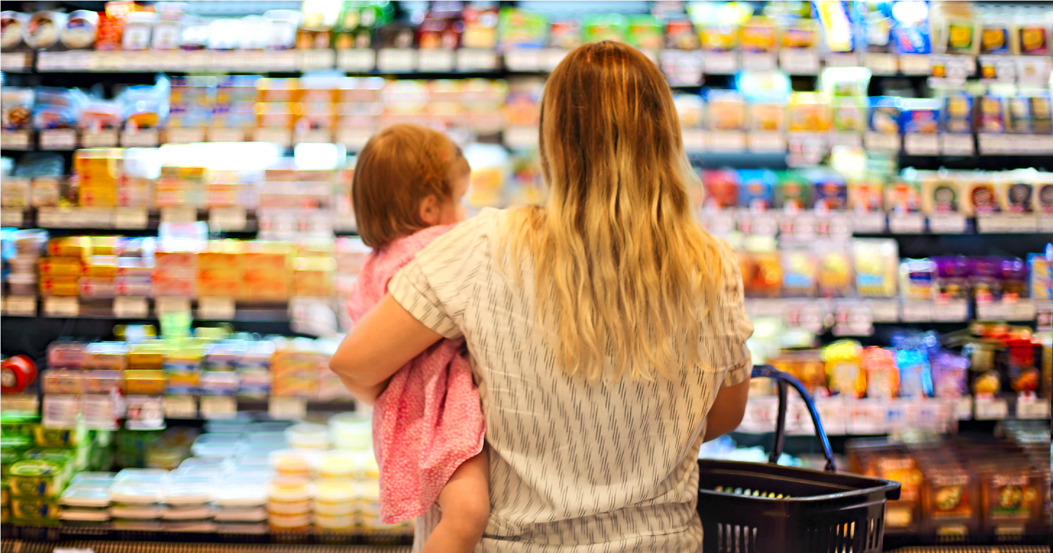 A woman looks over groceries with a child on her hip and her back to the camera.