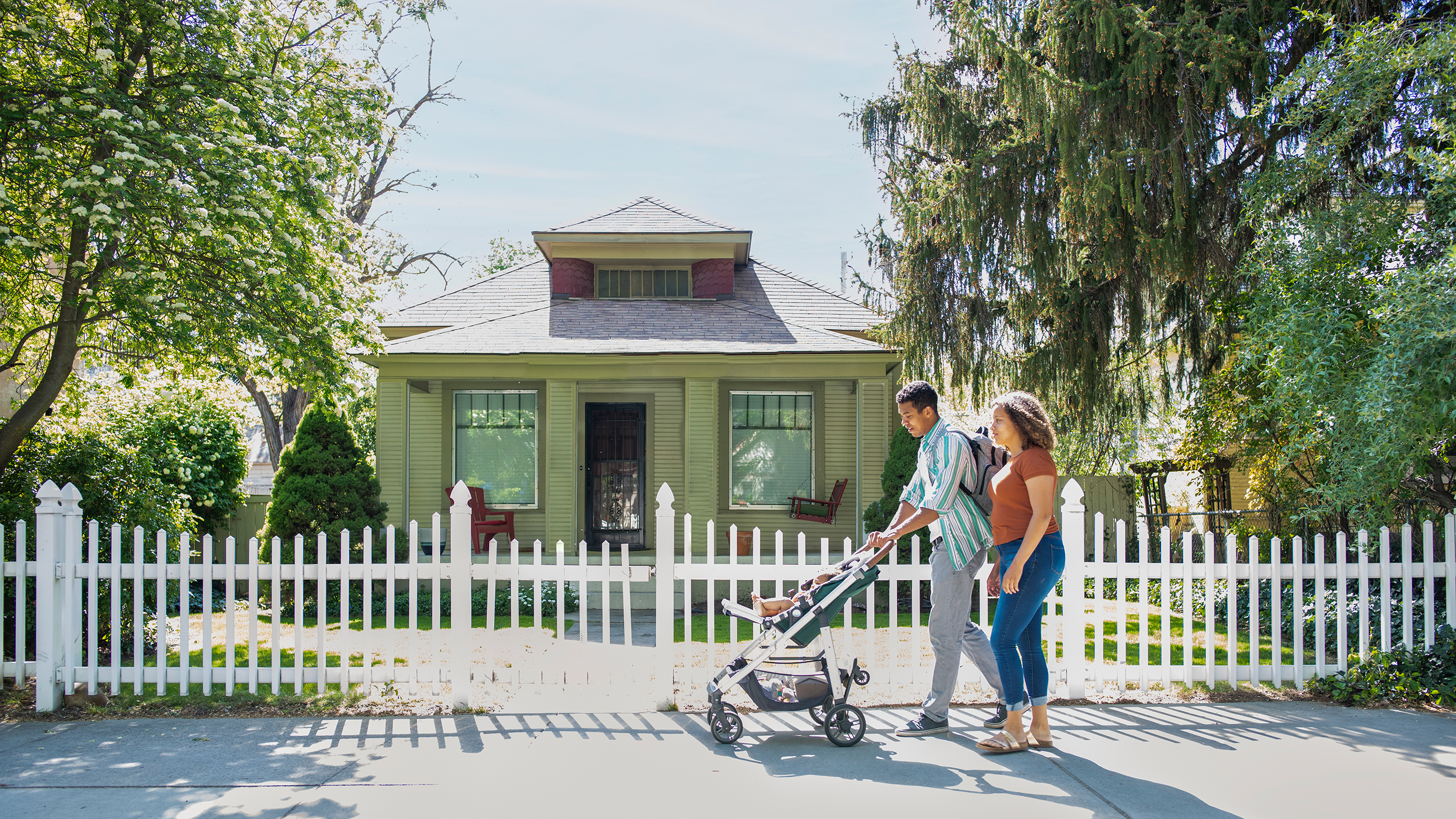 A mother and father pushing a baby in a stroller down a neighborhood sidewalk.