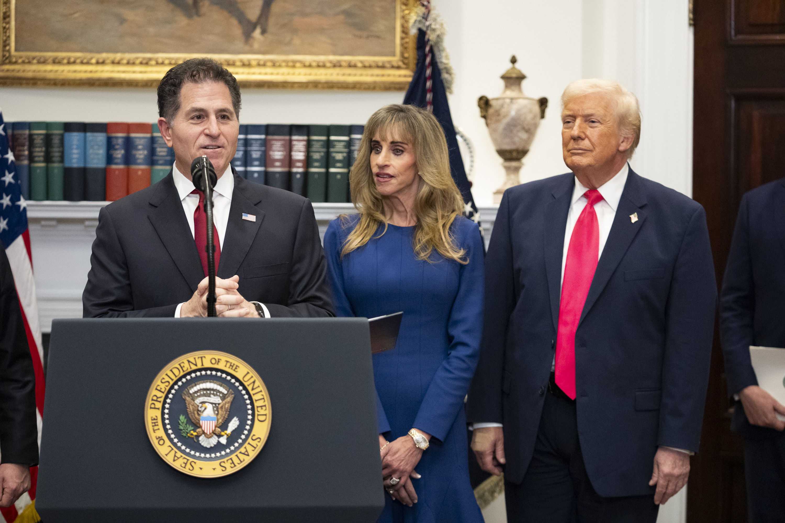 President Donald Trump and Susan Dell look on as Michael Dell speaks at a podium in the White House. 