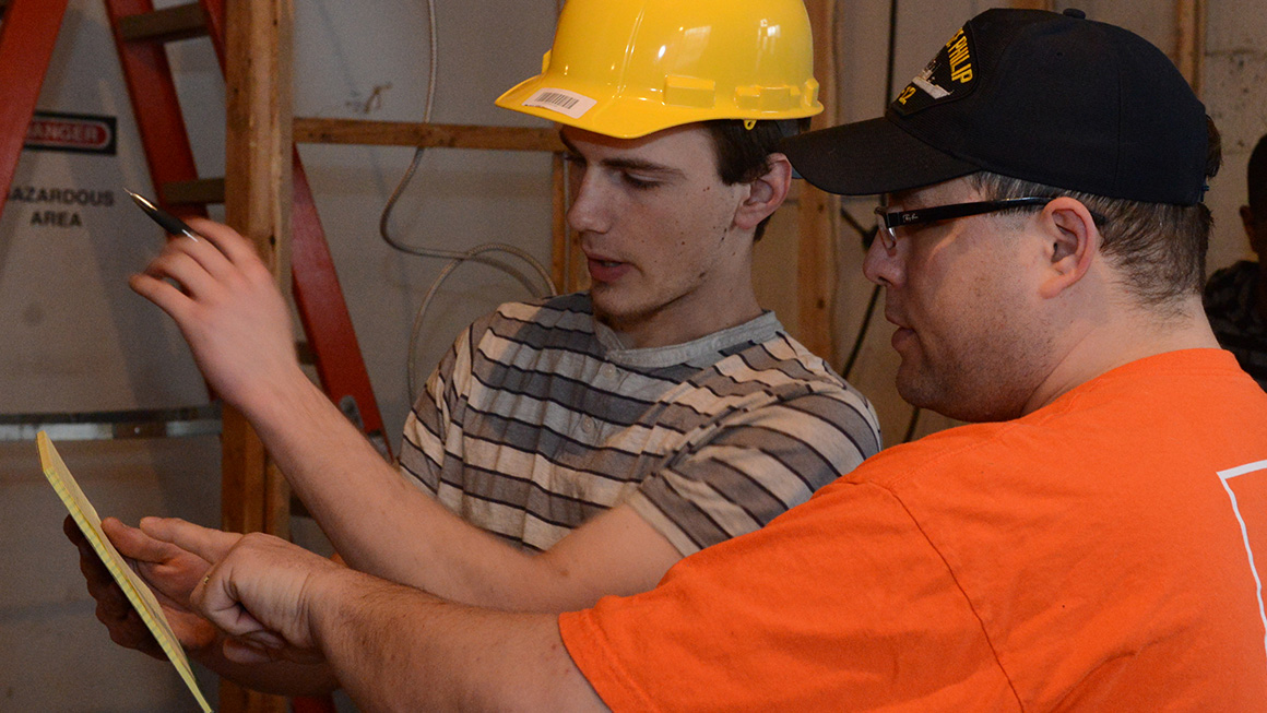 Two construction workers review a checklist inside a framed building, one pointing at a wall.