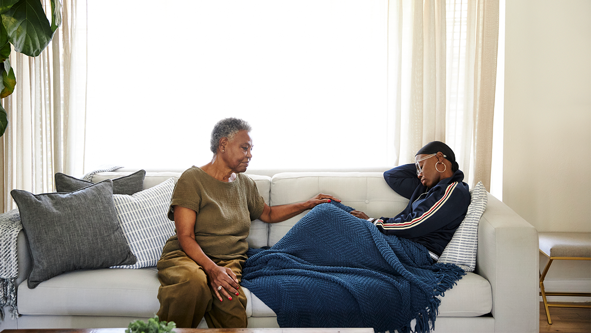 An older adult sits on a couch gently resting a hand on a younger person who is lying under a blue blanket
