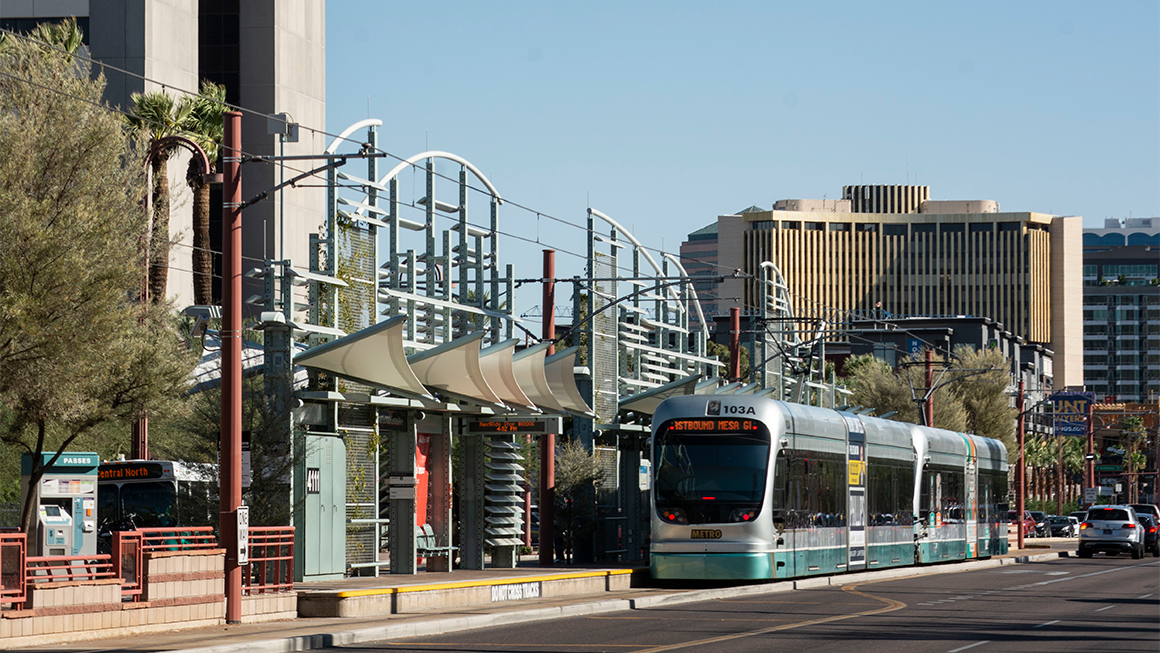 Light rail train arriving at a modern outdoor station with curved canopy structures and city buildings in the background.