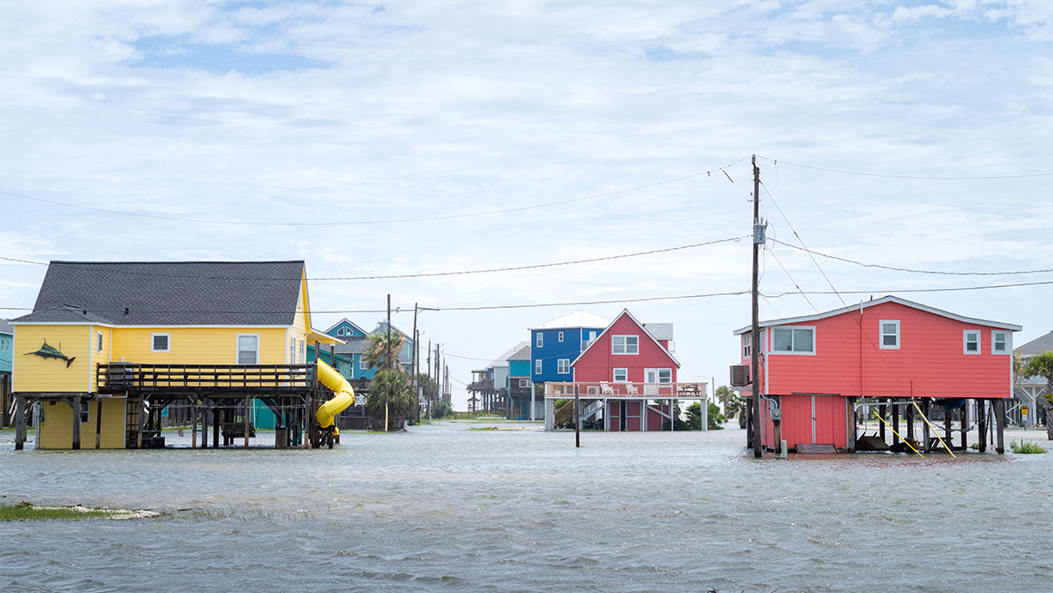 Floodwaters surround colorful stilt houses in a coastal neighborhood, with water covering streets beneath an overcast sky.