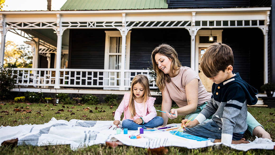 Photo of a mother and two young kids painting on the grass in front of their rural home. 