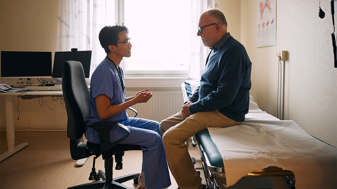 Health care professional in scrubs sits facing a patient on an exam table in a medical examination room, speaking during a consultation.