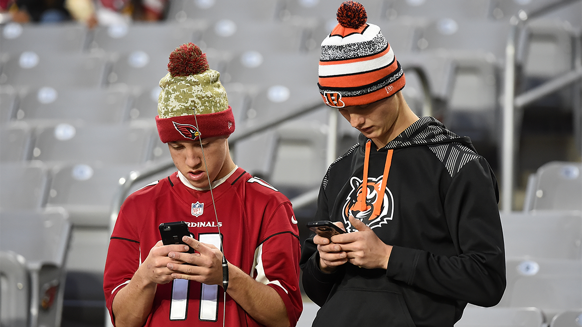 Two sports fans in team beanies and jerseys stand in stadium seating, looking down at smartphones.