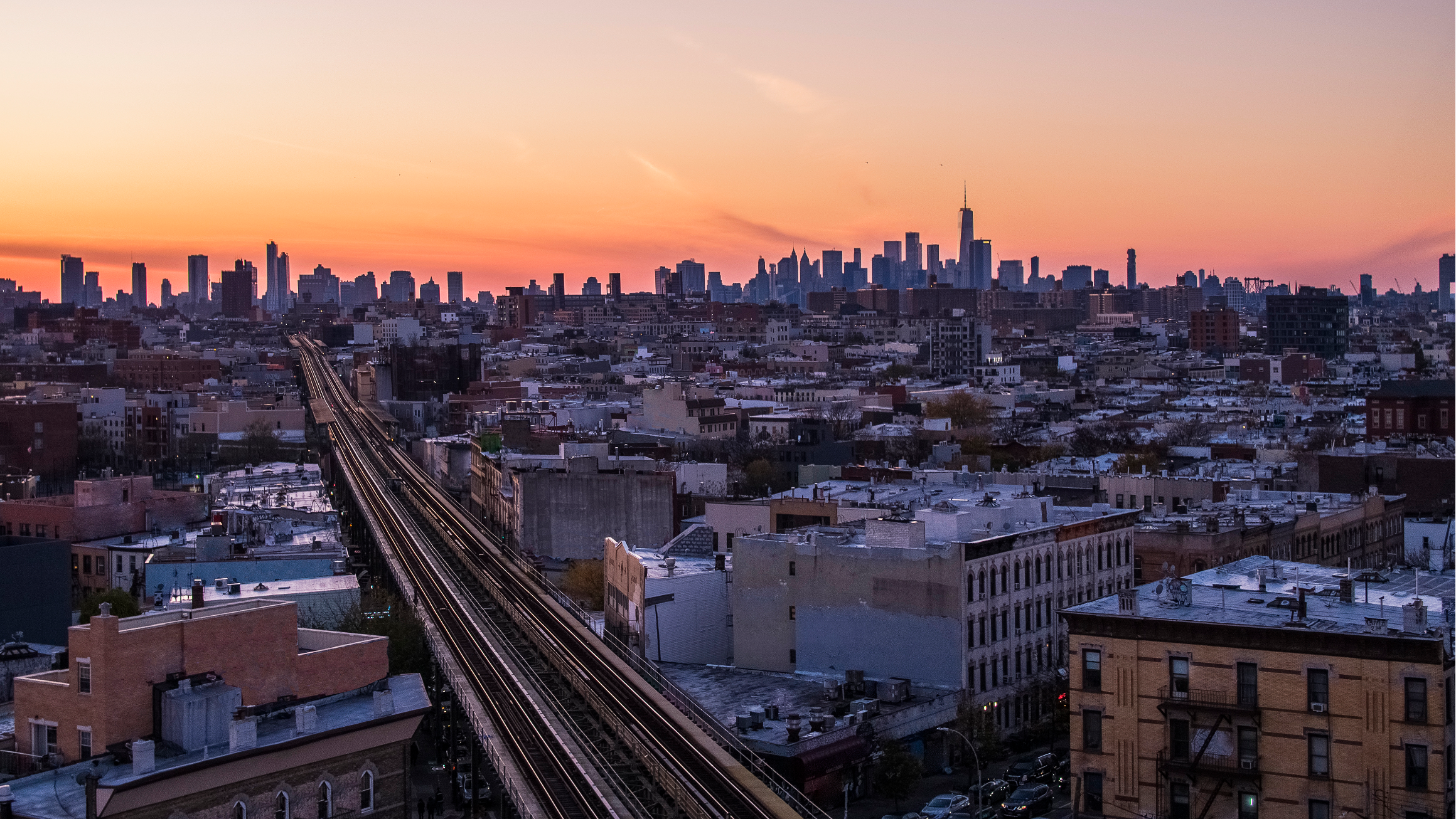 Panoramic city skyline at sunset, with elevated train tracks leading through low-rise buildings toward a cluster of downtown skyscrapers under a pink-orange sky.