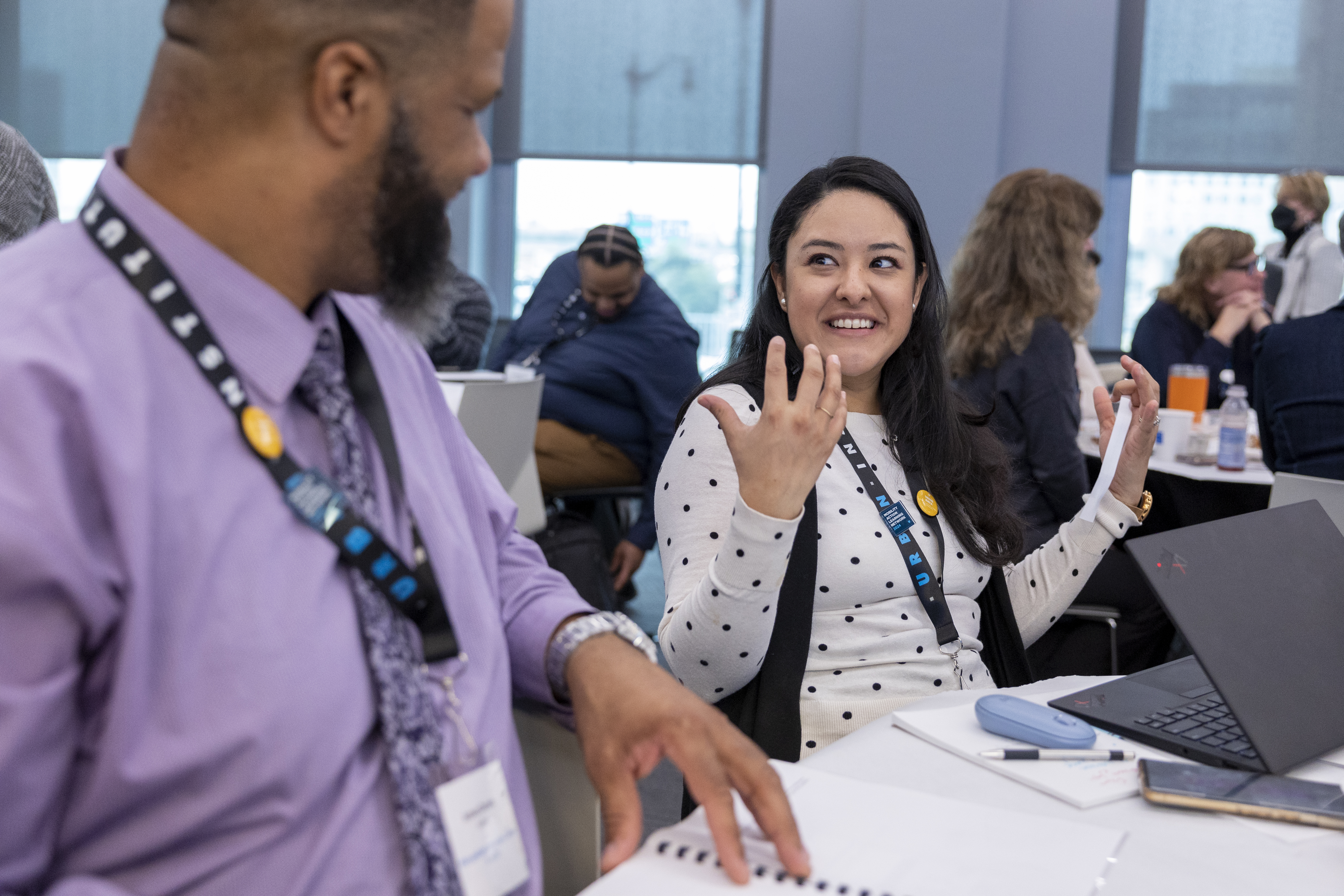 Two attendees wearing conference lanyards sit at a table during a workshop or meeting. One attendee gestures while speaking to another attendee in the foreground. A laptop, notebook, pen, phone, and other participants are visible in the bright conference room.