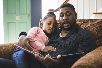 A black father reading to his daughter on their living room sofa. 