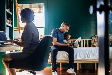 Father reading to baby as mother is on the computer.