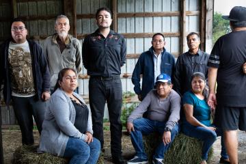 Víctor María Chamán, at center in a black button down shirt with an embroidered floral pattern, stands resolutely in a barn with eight storytellers and their families who stand next time him or sit on hay bales.