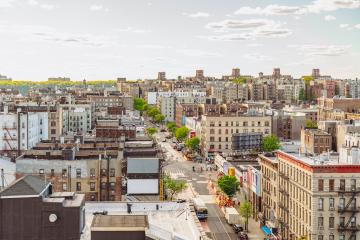 An elevated vantage point shows cars, busses, and people navigatinga street going through an urban setting dense with buildings and some trees.