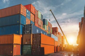 A tall crane extends over a wide and long stack of green, blue, and orange shipping containers as the sun rises.