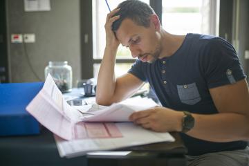 A man looks concerned as he looks through a stack of bills and other mail, with his head in his hand.