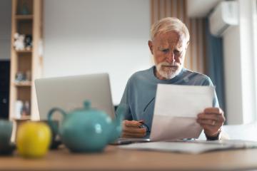 A bearded senior man sits at a table with his laptop in front of him, looking concernedly at a document.