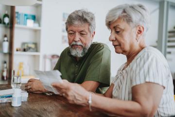 An elderly man and woman sit at their kitchen table analyzing a medical bill.