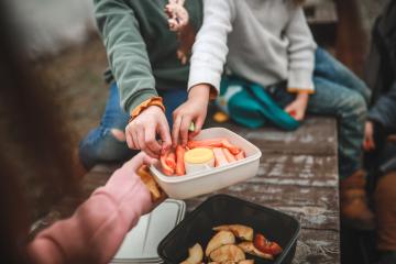 Two childen in sweaters reach for carrot sticks in a portable lunch container that's being held out by a third person, with whom they share a bench outside.