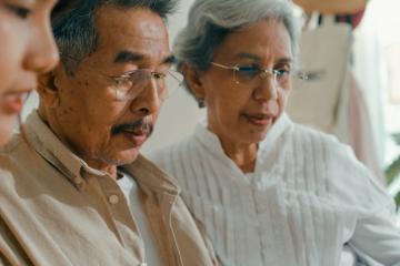 An elderly man and woman inspect something off camera, maybe something on a desk, along with a younger woman who appears to be explaining or helping.
