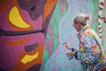 A blonde woman in a colorful jacket paints a multicolored mural on an outside wall.