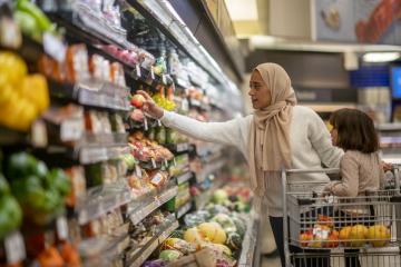 A woman pushing a grocery cart down a store's produce aisle reaches for an apple, with her little girl seated at the front of the cart and some oranges in the basket.