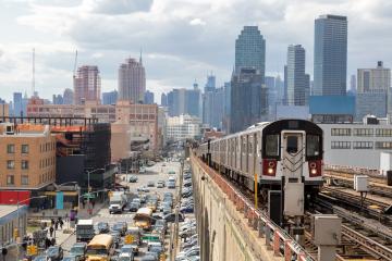 A busy urban skyline on a cloudy day, with lots  of people walking and cars visible on the street and parked, with a public train heading toward the camera.