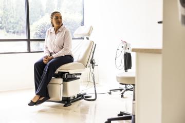An anxious-looking woman in blue slacks and a pale pink button-down shirt sits with her hands clasped together on an exam chair in a doctor's office.