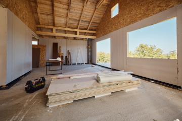 Tools and wooden panels sit on the ground of a home under construction on a sunny day, and other construction materials line the far wall under exposed beams.