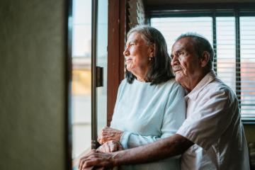An elderly man embraces an elderly woman as they look out a window, presumably in their home.