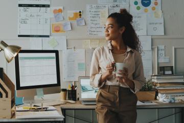 A young woman in a business-casual outfit holds a coffee mug, looking out a window to her right, standing in front of her desk, which features plenty of evidence of productivity: a calendar, a stack of books, motivational quotes, a desk top, a laptop, and even a backup cup of coffee.
