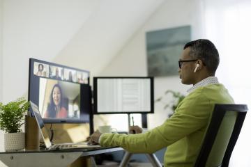 A man wearing a business casual shirt and sweater sits in front of a desk with a laptop and two additional monitors, holding a virtual meeting with colleagues.