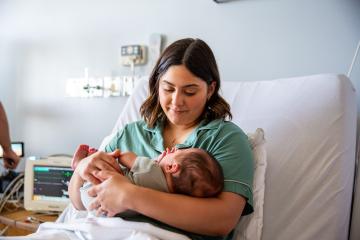 A young mother reclined in a hospital bed smiles down at her baby, as a person in the background adjusts medical equipment.