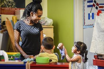 A child care provider smiles at two toddlers in a sunny classroom