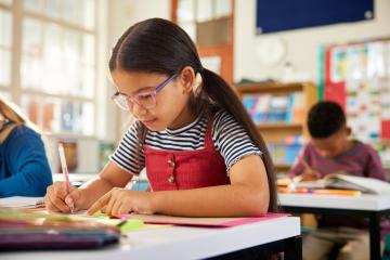 A young girl of color with long pigtails, freckles, and pink glasses uses an orange gel pen to do her schooldwork in a pink folder.