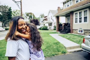  A happy little girl hugs her mom, who is smiling so hard her eyes are closed and her nose is crinkling, stand on the driveway in front of a two-story home.
