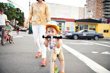  A little boy wearing a colorful helmet and riding a colorful scooter smiles behind his handlebars as he crosses a city street with his mom right behind him, followed by a brother on a bike with training wheels and their father.