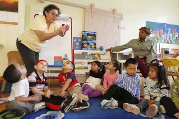 A class of small children sit in a circle in a classroom as the teacher in a yellow shirt and glasses presents on a strawberry, with the teacher's aid sittings nearby, looking at a diagram of the solar system.