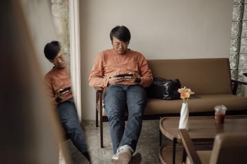 A man in a long-sleeve orange t-shirt and blue jeans man sits on a brown leather bench  while looking intently at his cell phone.