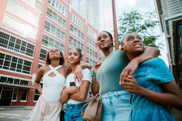 A Black mother stands with her three children, her arm around the child on her left, and all four are standing alongside a city street while smiling at something off-camera. 