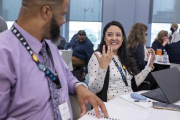 Two attendees wearing conference lanyards sit at a table during a workshop or meeting. One attendee gestures while speaking to another attendee in the foreground. A laptop, notebook, pen, phone, and other participants are visible in the bright conference room.