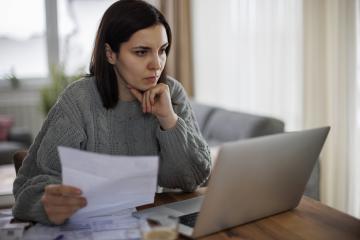 A woman with short brown hair and a sage-colored cable sweater sits at a countertop, holding a bill with one hand while supporting her chin with the other and looking at her laptop with a furrowed brow.