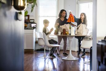 A family unpacks groceries at the kitchen table. A young girl holds up a bag of oranges.