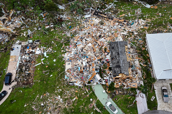 Aerial view of a neighborhood showing severe storm damage, with one home reduced to debris and wreckage scattered across surrounding yards.