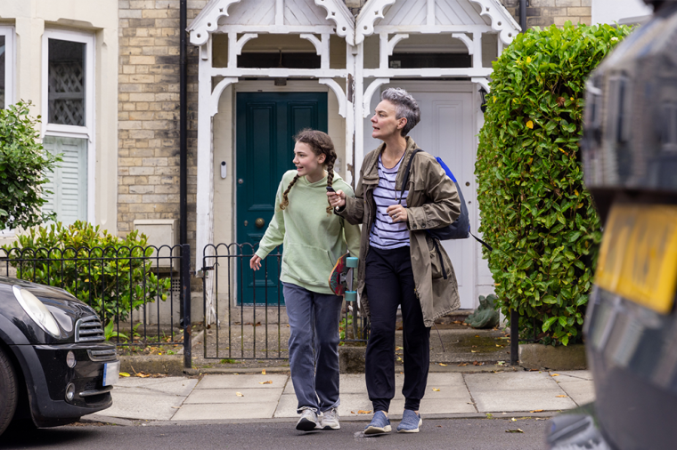 A parent with a backpack and child with a skateboard cross the road on the way to school.