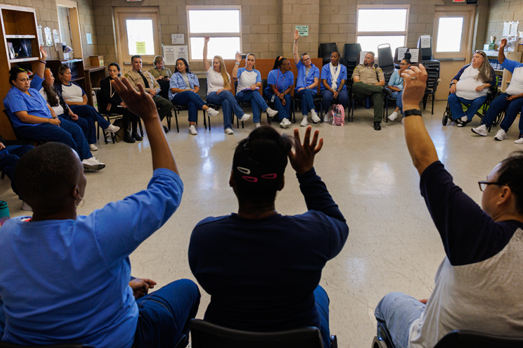 About two dozen incarcerated women sit in chairs arranged in a circle in a classroom. Some raise their hands.  