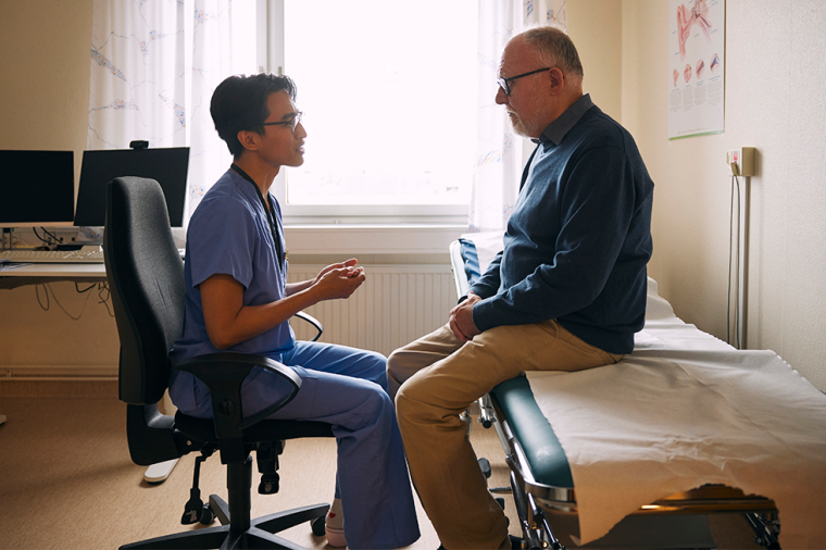 Health care professional in scrubs sits facing a patient on an exam table in a medical examination room, speaking during a consultation.