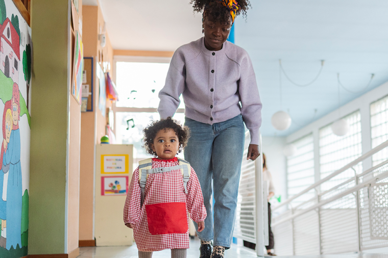 A young child walking through the halls of a day care, followed by her mother.